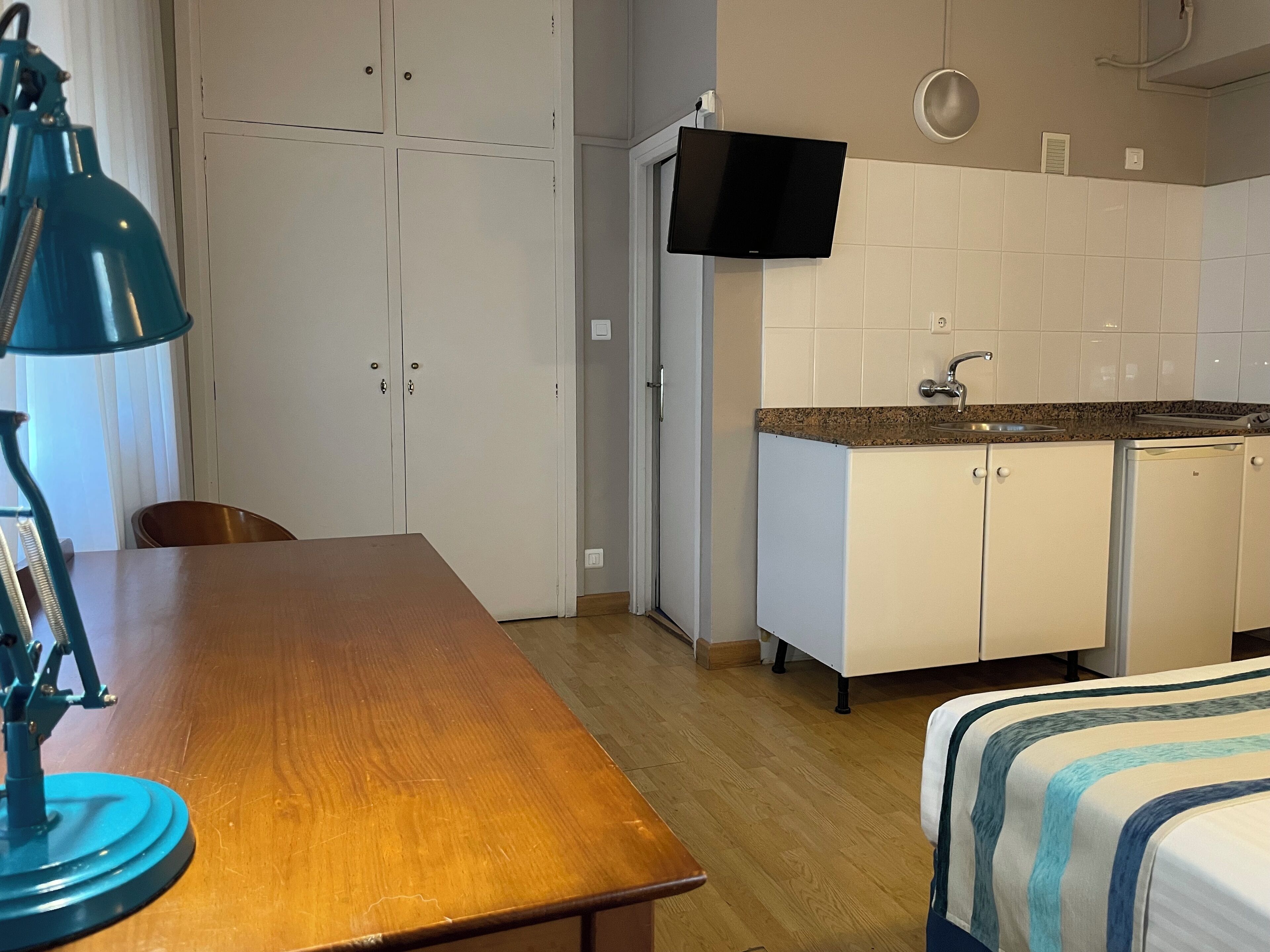 Una stanza con un tavolo di legno, una lampada blu, un letto con lenzuola a righe, un angolo cottura con pensili bianchi e una TV montata sopra il bancone.