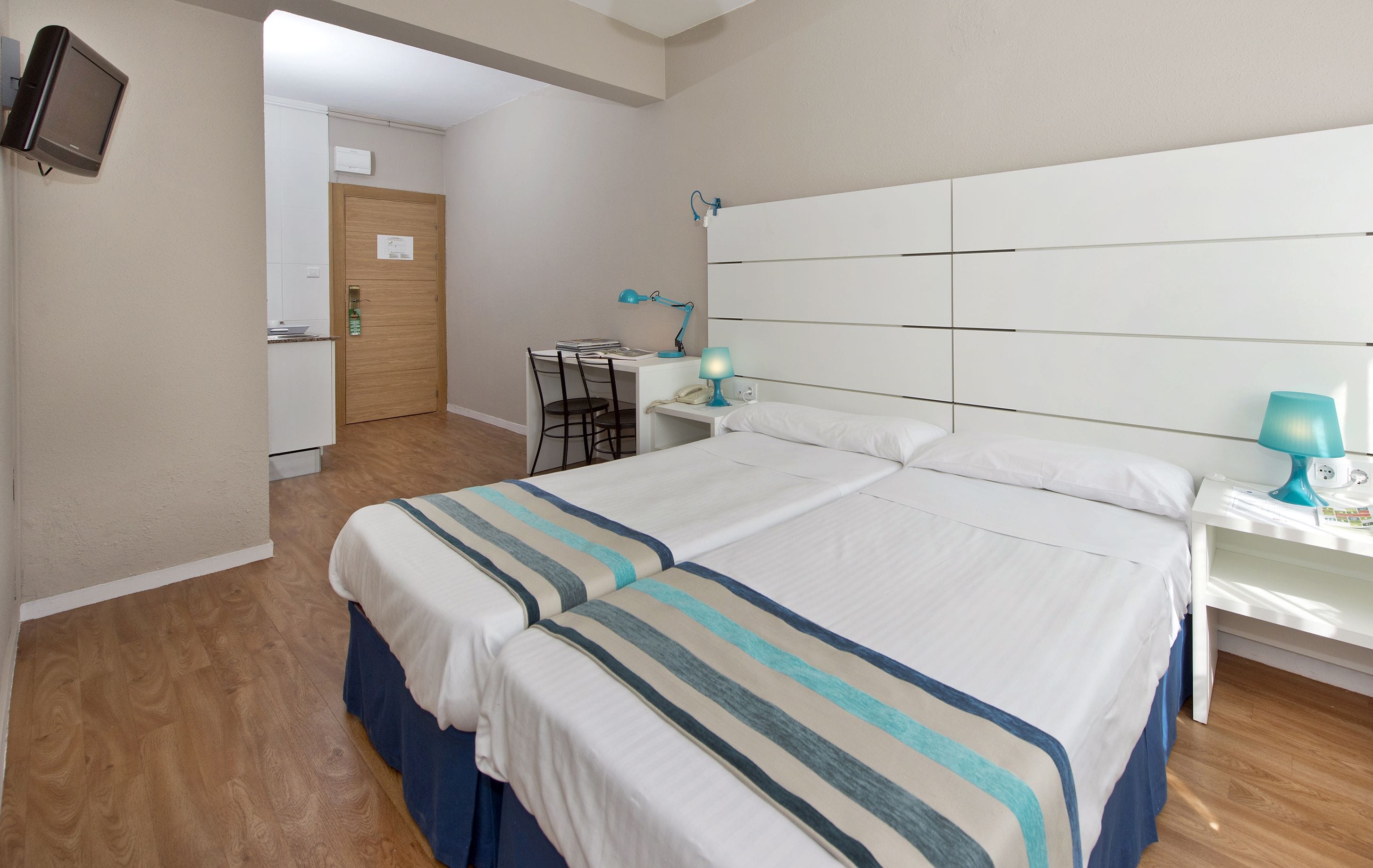 A hotel room with a large bed, a desk with a chair, a small table with a lamp, and a television mounted on the wall.