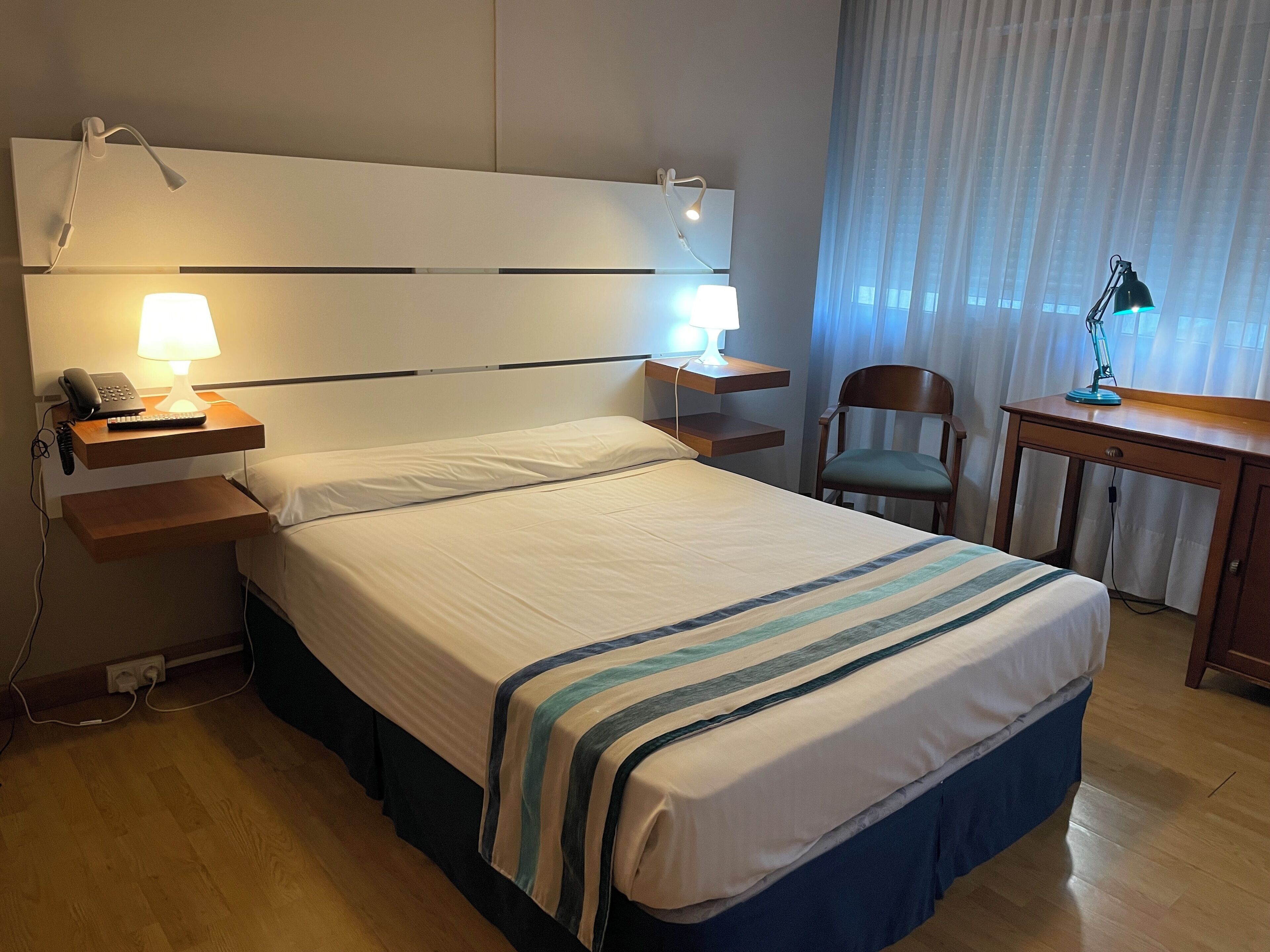 Habitación de hotel con una cama grande, lámparas de noche, un escritorio con silla y una ventana con cortinas.