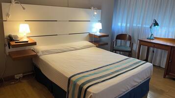 Habitación de hotel con una cama grande, lámparas de noche, un escritorio con silla y una ventana con cortinas.