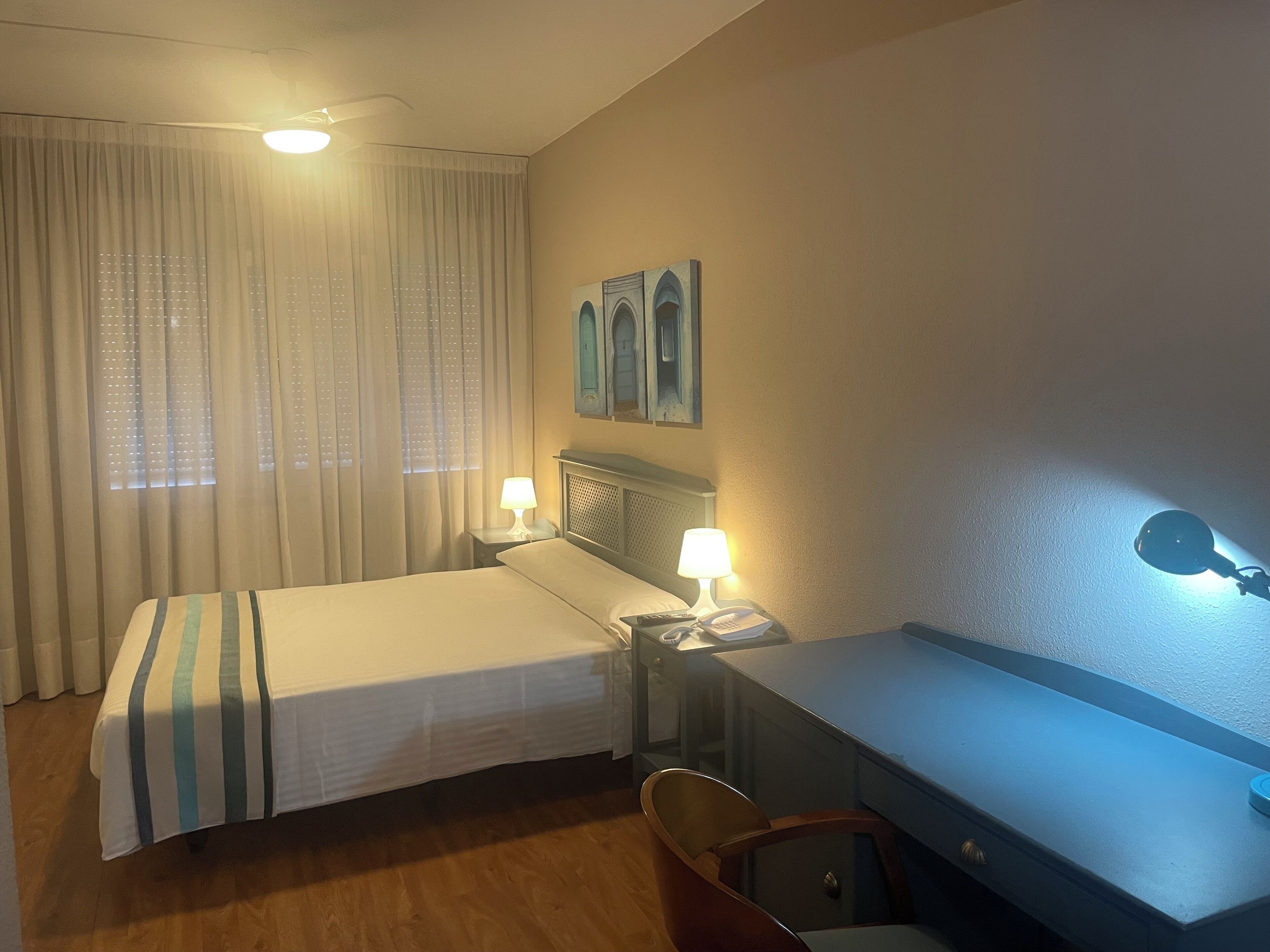 A hotel room with a bed, a desk, a chair, a lamp, and a ceiling fan.