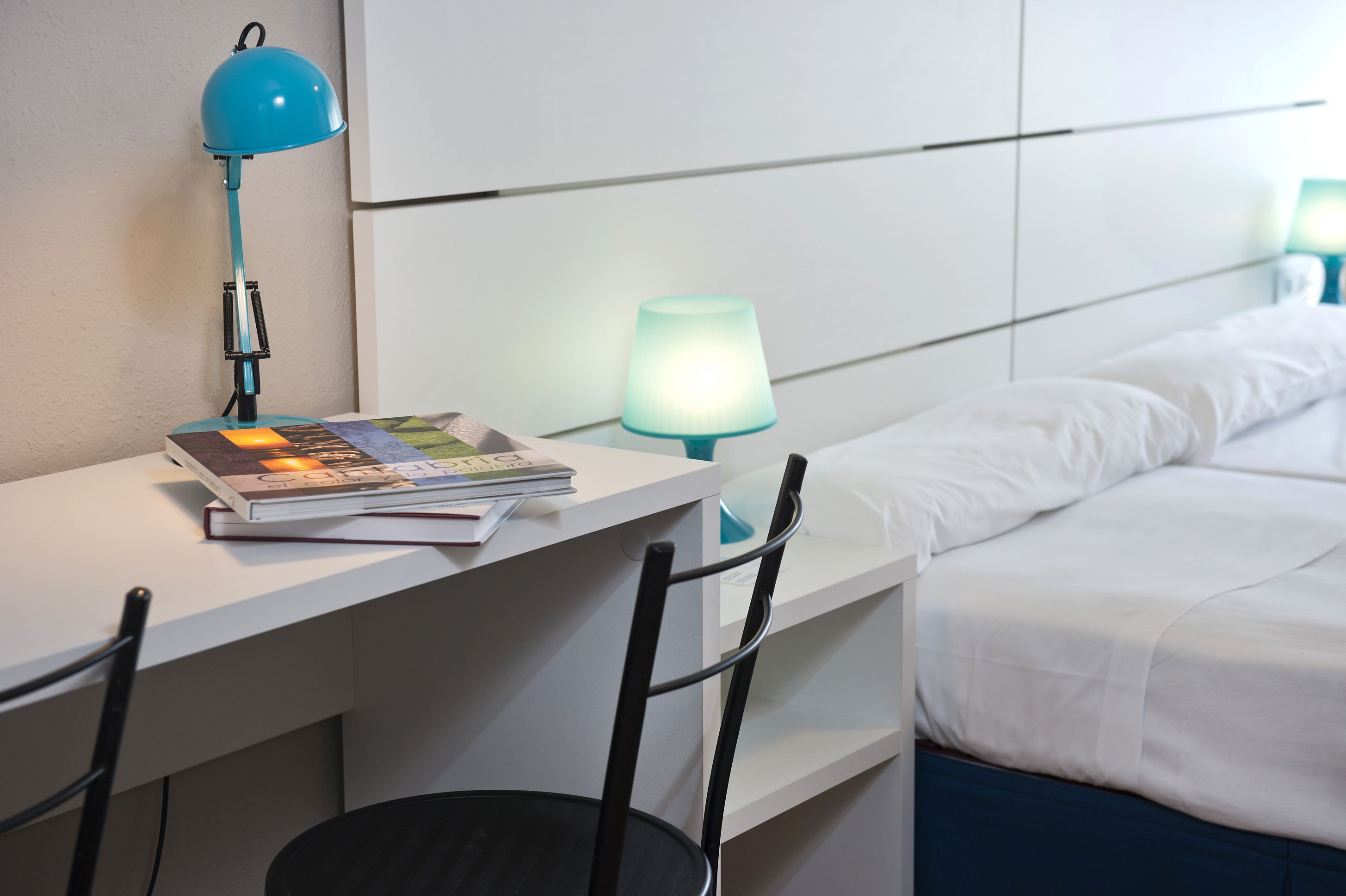 Et hotelværelse med et skrivebord, en stol, en seng med hvide sengetæpper og to sengeborde med lamper.