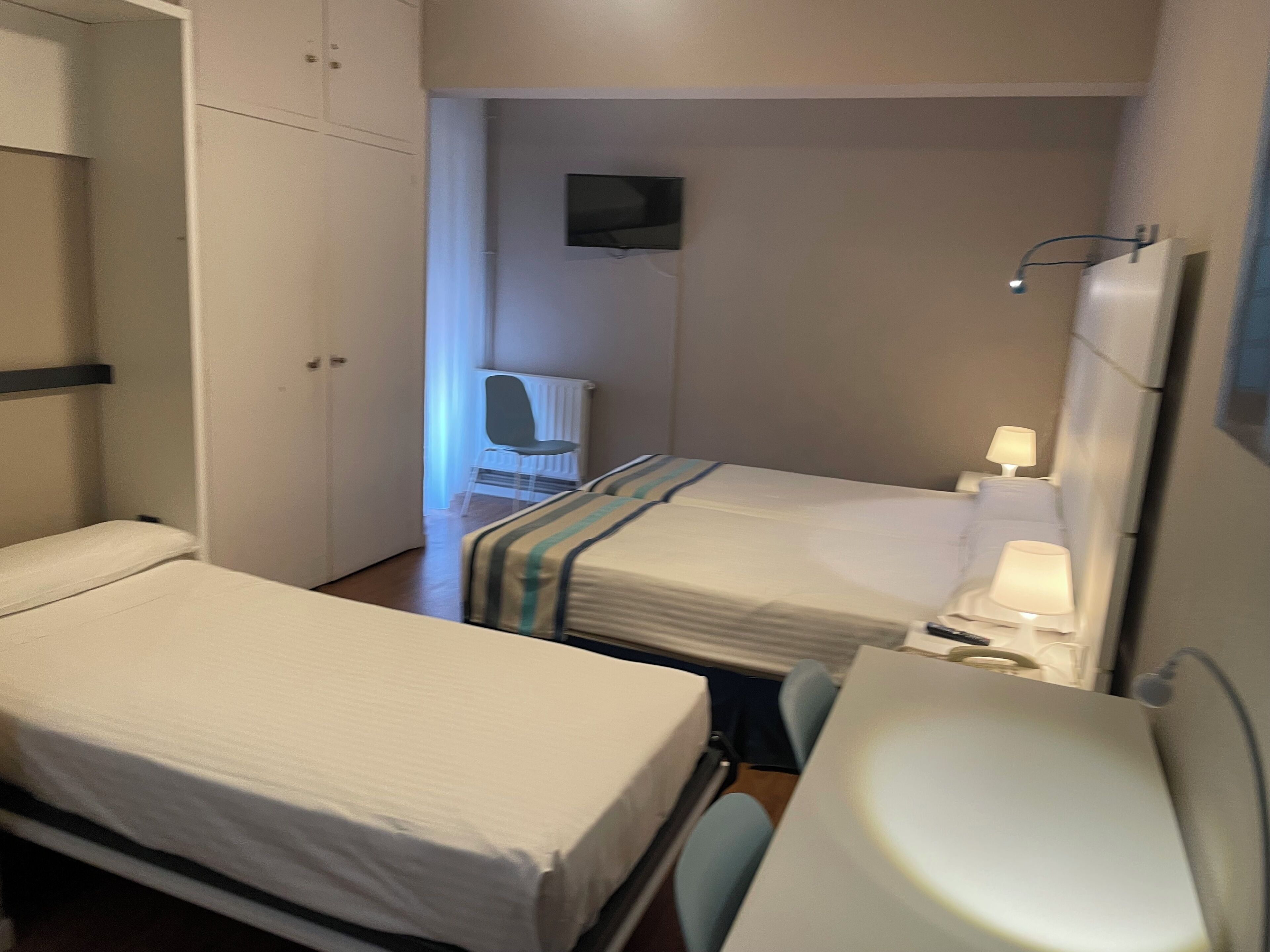 A small, compact hotel room with two single beds, a bedside table with a lamp, a TV mounted on the wall, and a radiator.