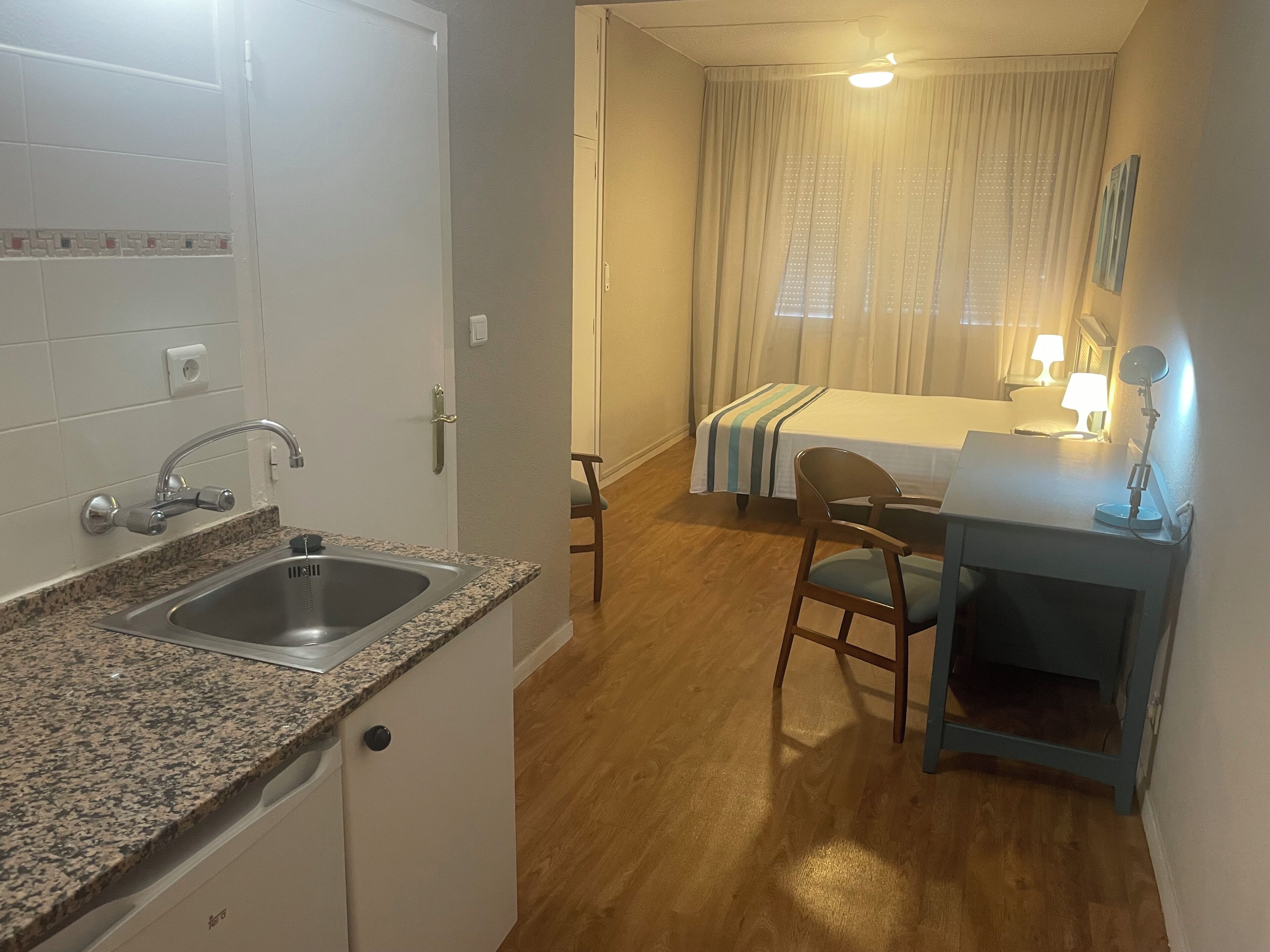 A compact hotel room with a kitchenette, a bed with a striped bedspread, a desk with a lamp, and a chair.