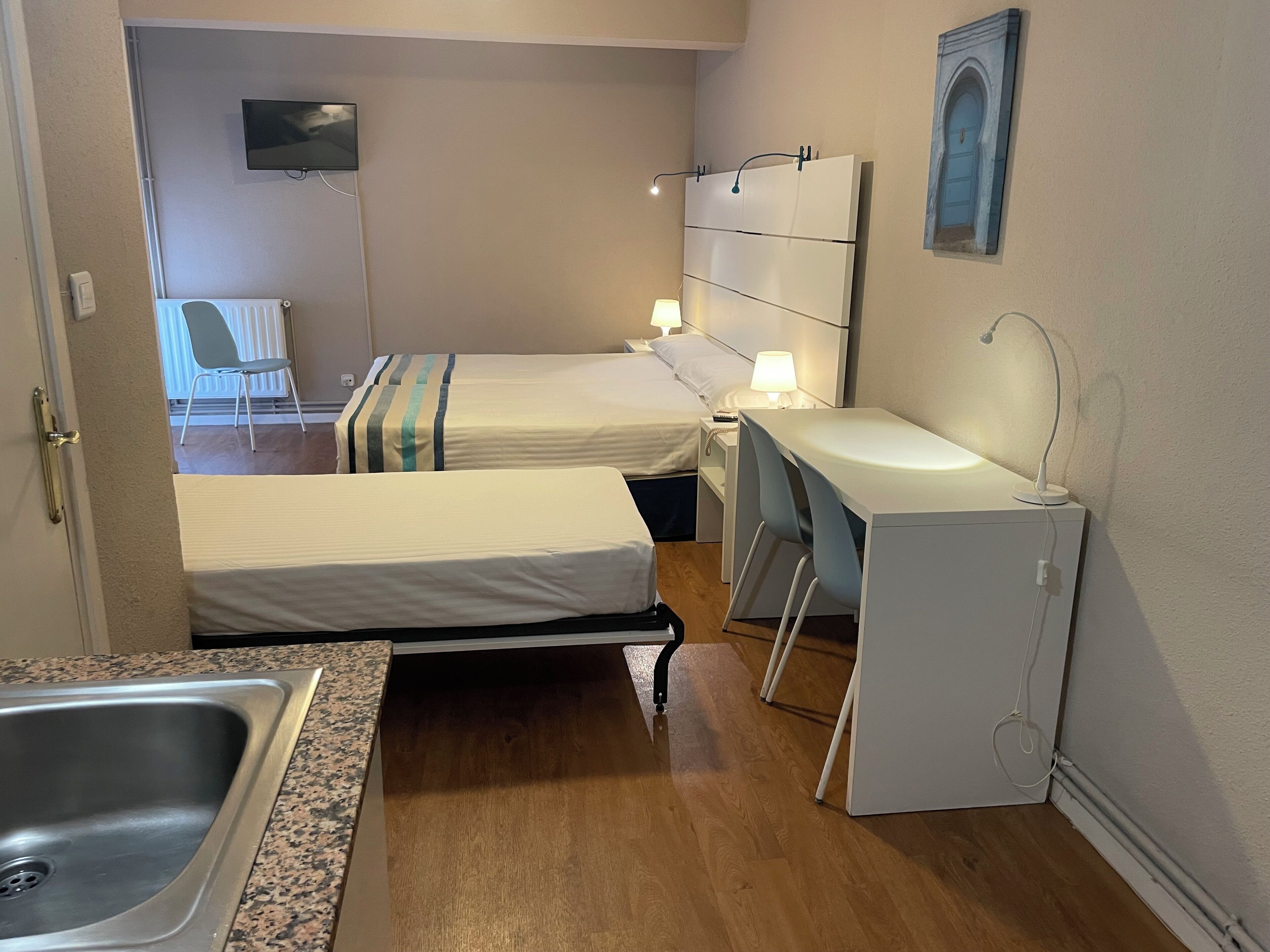 A compact hotel room with two beds, a small desk, a sink, and a TV mounted on the wall.