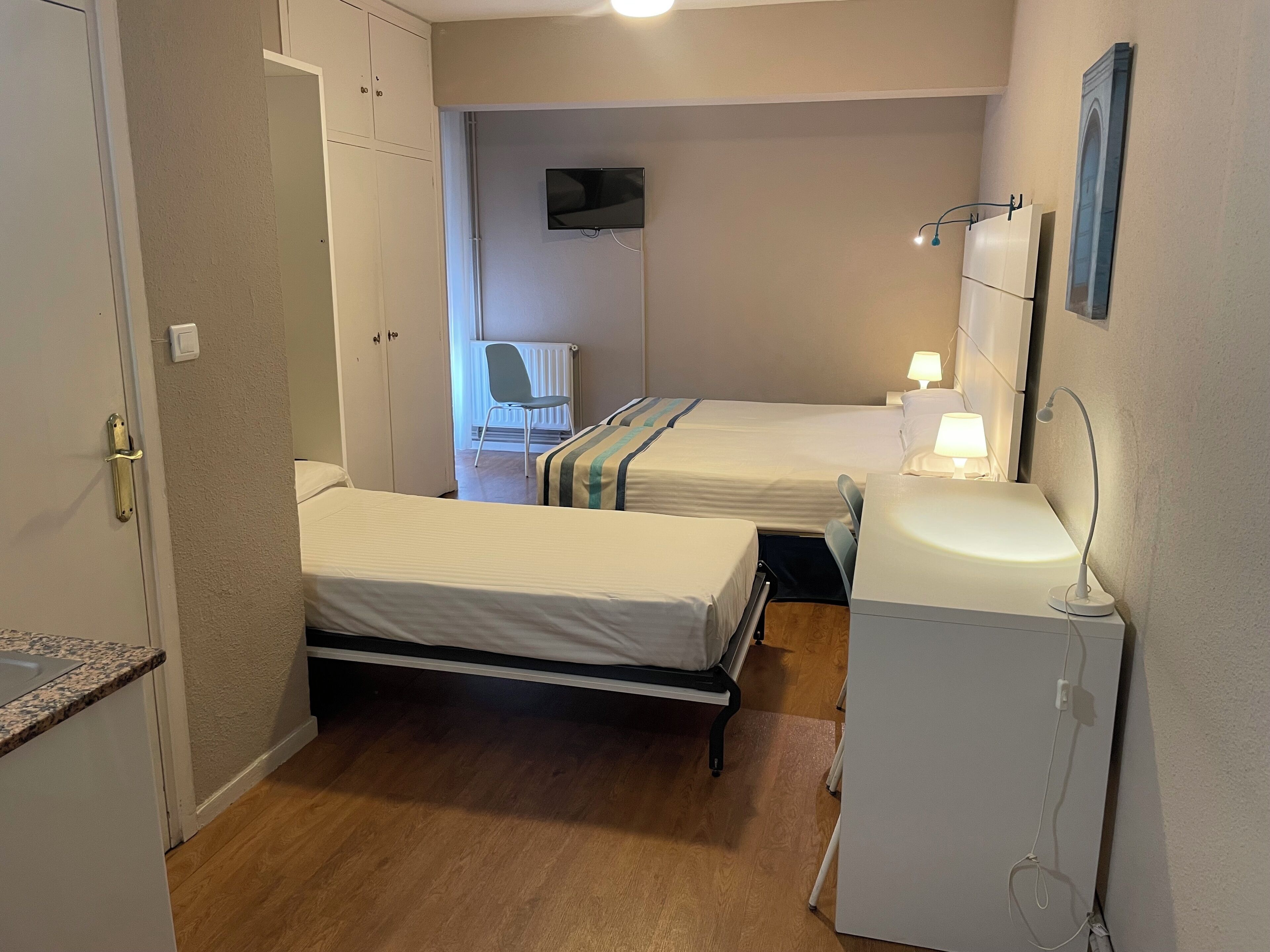 Ett litet, väl upplyst rum med två sängar, ett skrivbord, en stol, en tv och ett badrum med handfat och spegel.