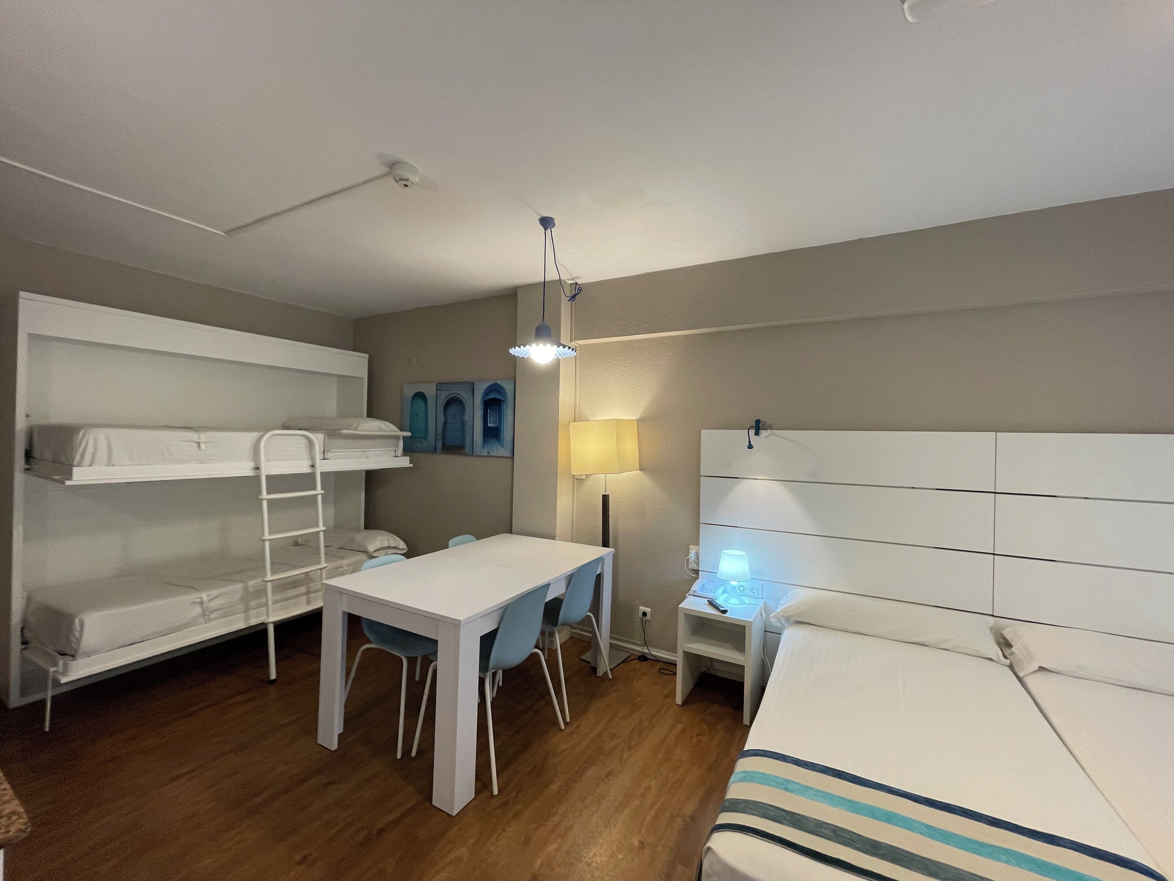 A dormitory room with bunk beds, a desk, a chair, a lamp, and a bedside table.