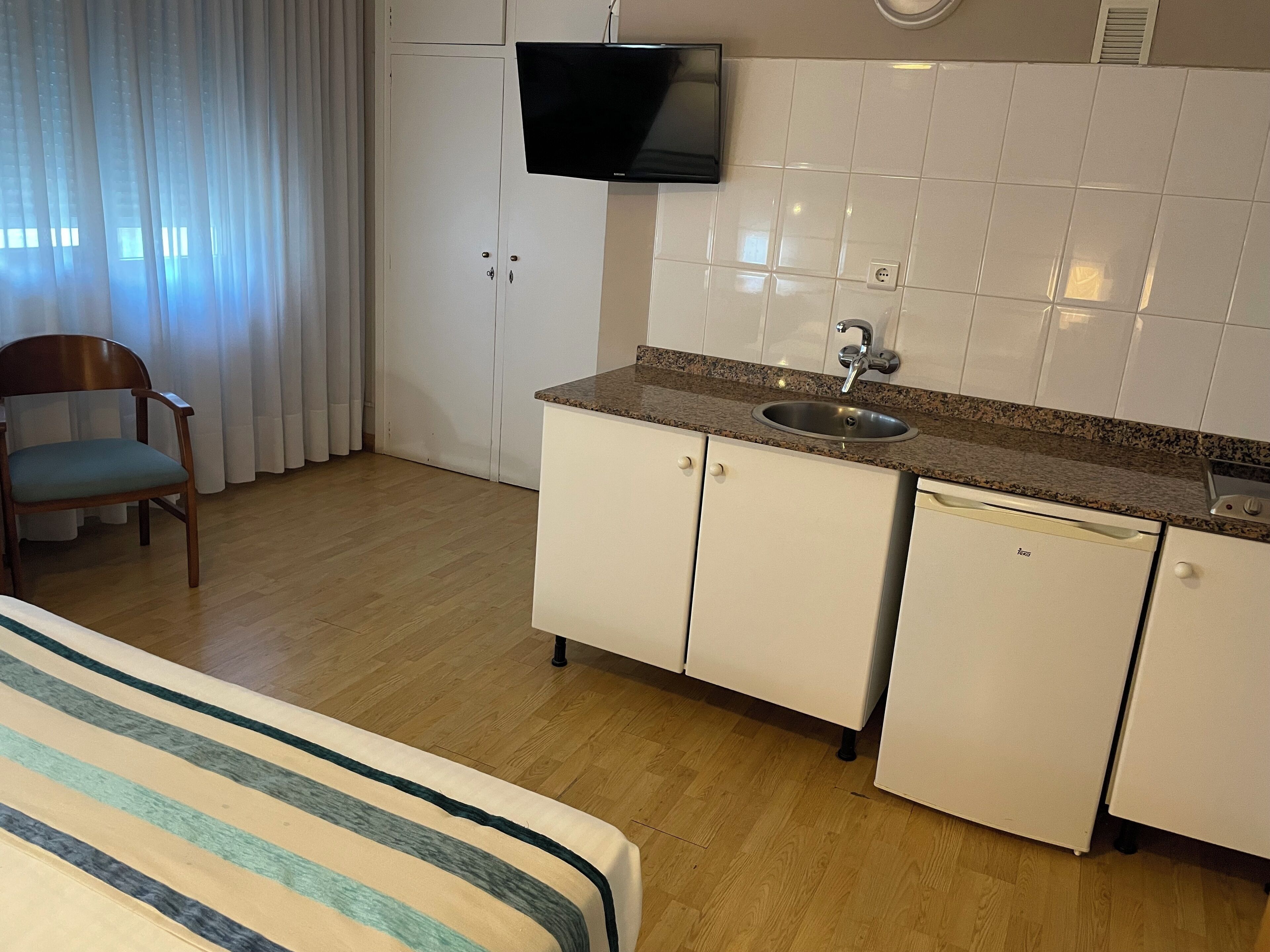 Quarto de hotel compacto com cama, bancada de cozinha, pia, televisão, cadeira e janela com cortinas.