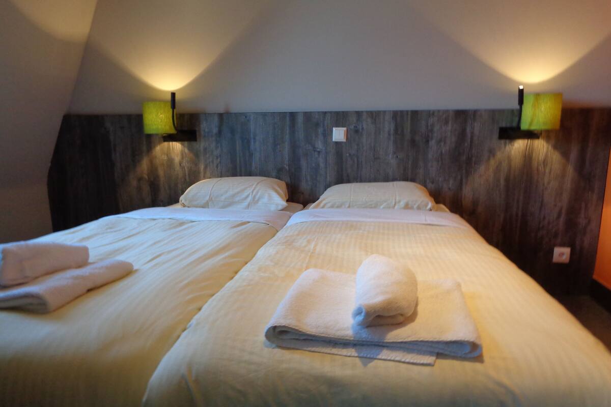 Een hotelkamer met twee bedden, elk voorzien van een netjes opgevouwen handdoek. De kamer heeft een houten hoofdbord en twee wandlampen.