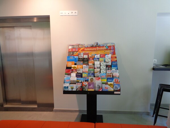 Una exhibición de folletos de viajes en el vestíbulo de un hotel, con un ascensor y una recepción al fondo.