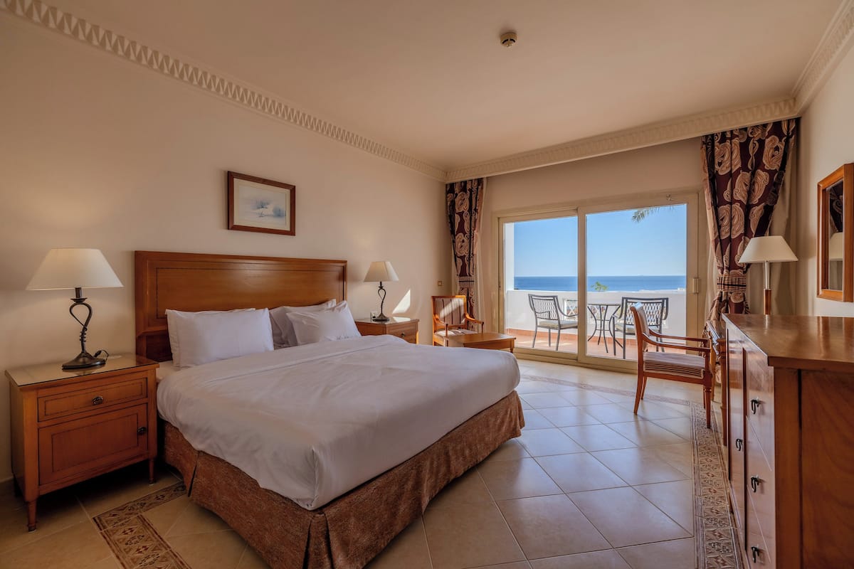 Habitación de hotel con una cama grande, dos mesitas de noche con lámparas, un cabecero de madera, un cuadro enmarcado en la pared y un balcón con vista al mar.