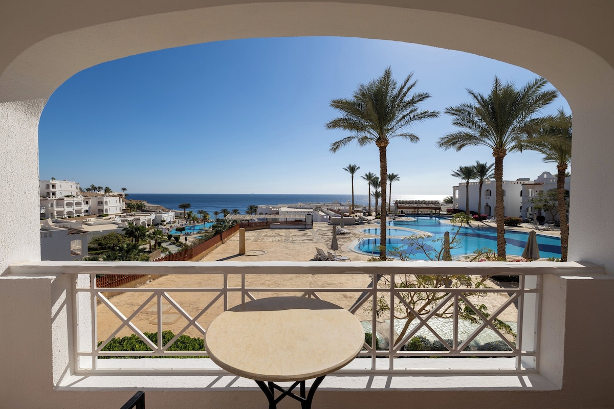 Un balcón con vista a una playa, una piscina y palmeras.