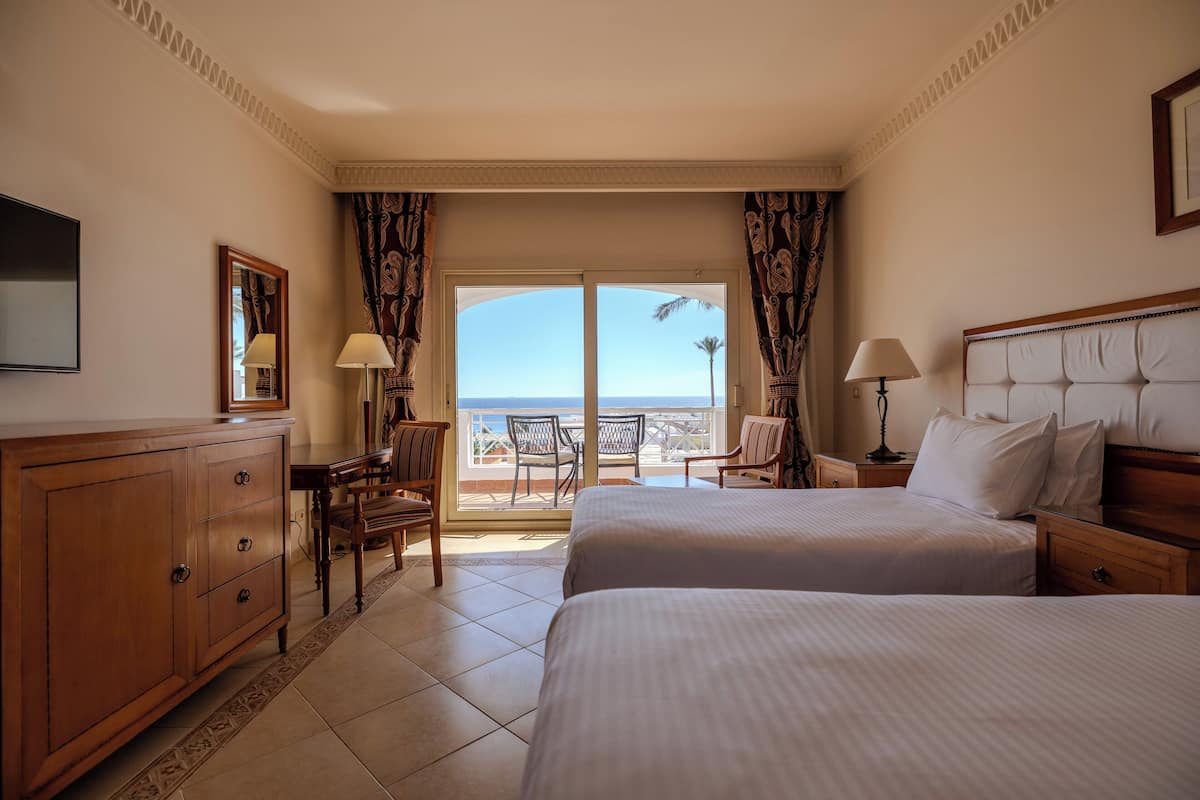 Habitación de hotel con dos camas, un aparador de madera, televisión y balcón con vista al mar.