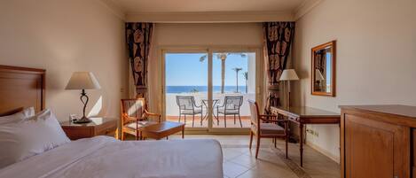 A hotel room with a large bed, a desk, a chair, and a view of the beach through the window.