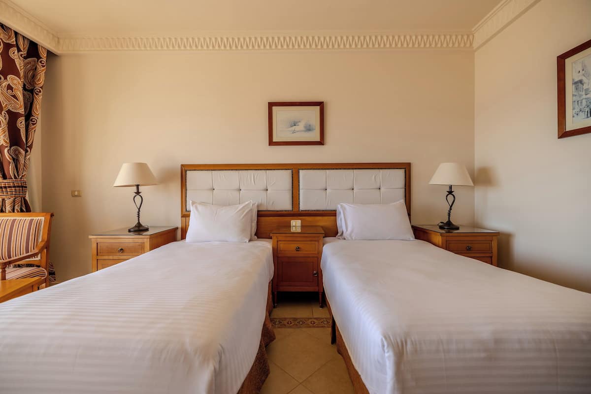 Habitación de hotel con dos camas individuales, cabecero de madera, mesitas de noche con lámparas, un pequeño armario y un cuadro enmarcado en la pared.