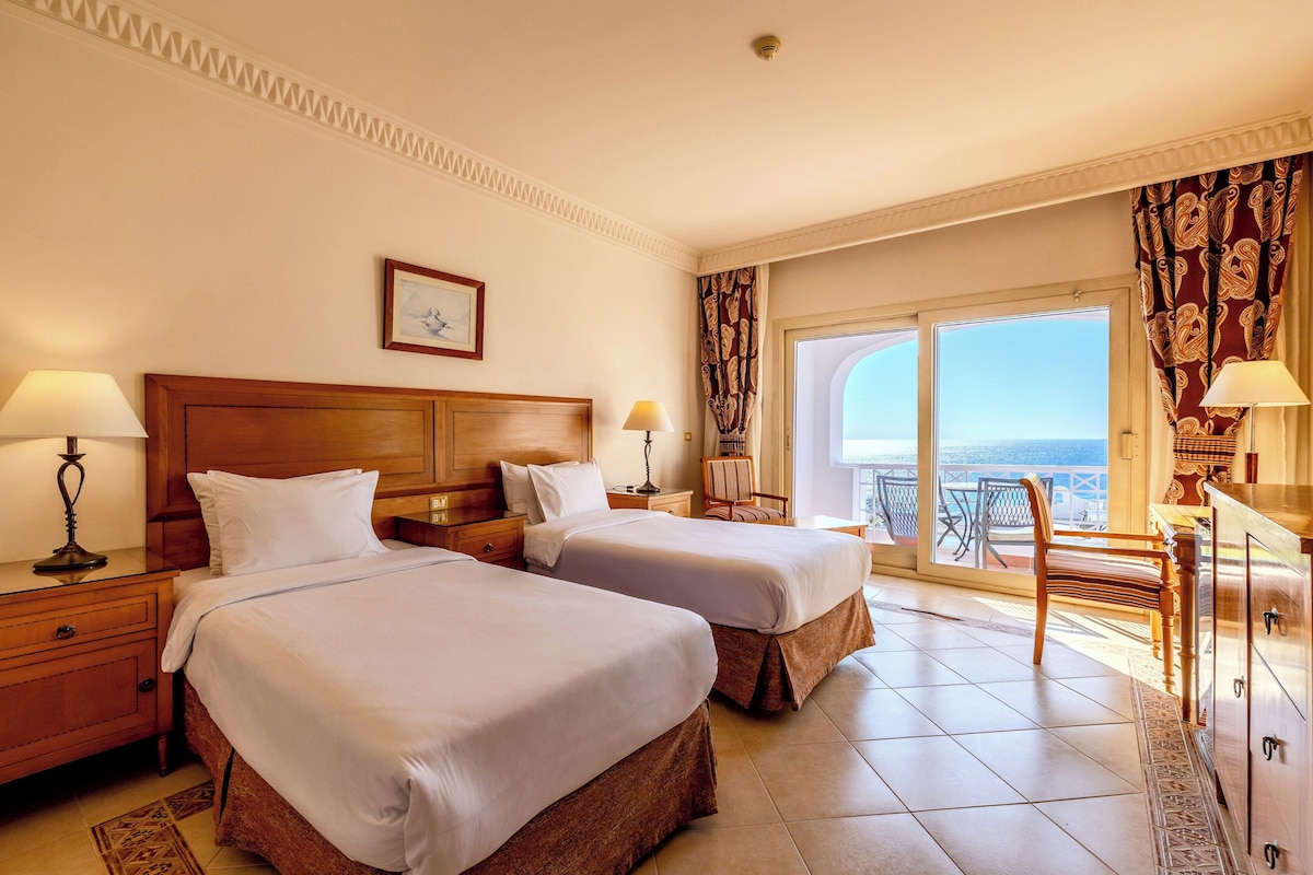 Habitación de hotel con dos camas, un balcón con vistas y un escritorio con lámpara.