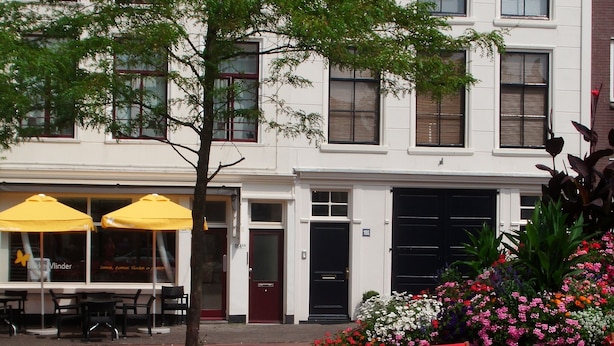 An outdoor dining area with red chairs and tables, a building with white walls and a red-tiled roof, and a flower bed with pink and white flowers. - Short Stay Maff Apartment (The Hague)