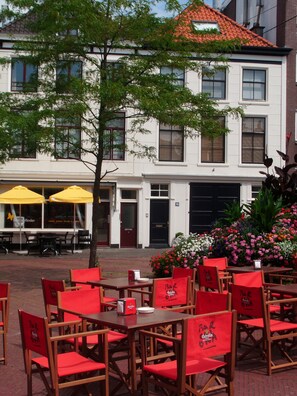 An outdoor dining area with red chairs and tables, a building with white walls and a red-tiled roof, and a flower bed with pink and white flowers. - Short Stay Maff Apartment (The Hague)