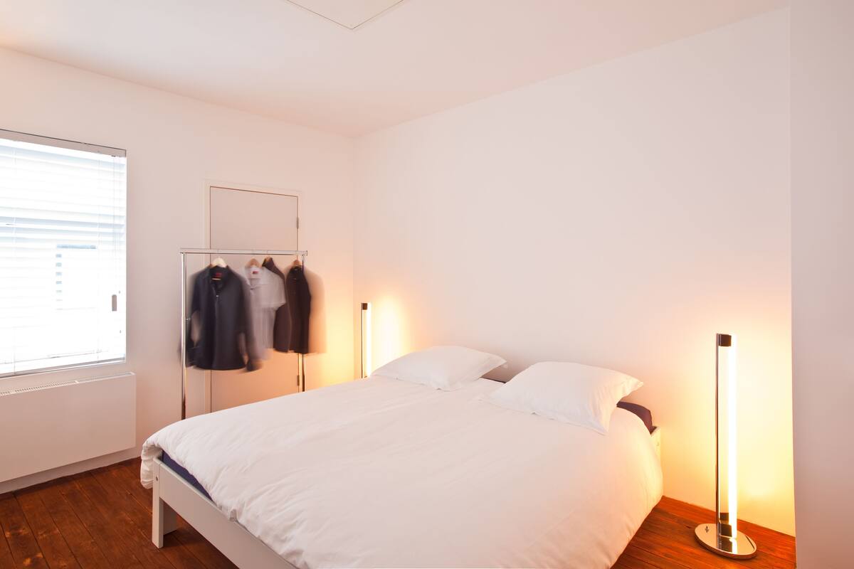 A bedroom with a bed, a nightstand, a bedside lamp, a mirror, and a window with blinds.
