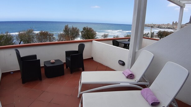 A balcony with a view of the sea, featuring two black wicker chairs, a small table, and a white chair with a purple towel. - Villa Cerniglia (Cefalù)