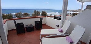 Un balcón con vistas al mar, que incluye dos sillas de mimbre negras, una mesita y una silla blanca con una toalla morada.