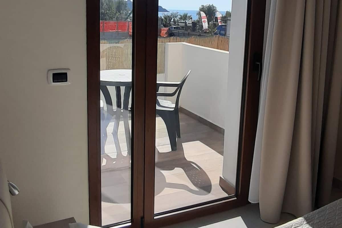 Habitación de hotel con cama, ventana con cortinas y vista a un balcón con mobiliario exterior.