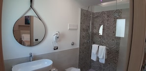 A modern bathroom with a round mirror, a white sink, a glass-enclosed shower, and patterned tiles. - Villa Cerniglia (Cefalù)