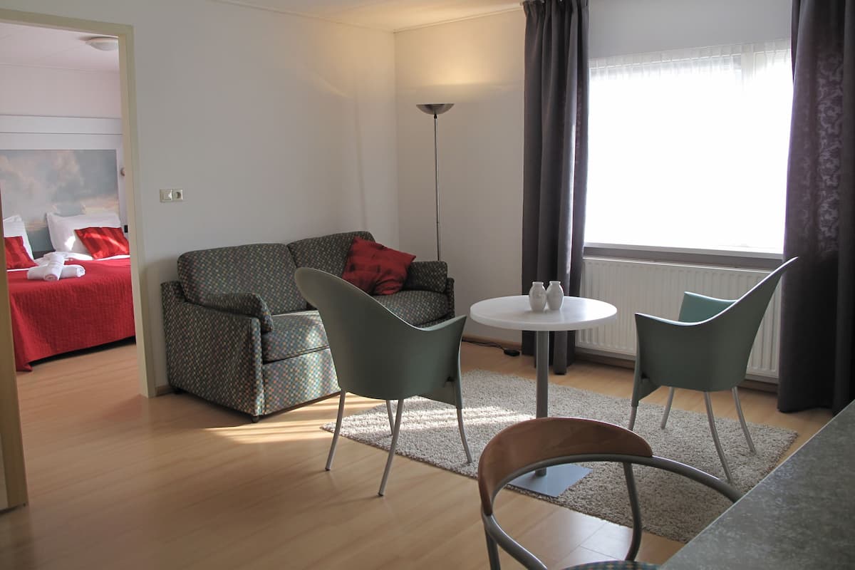 A modern living room with a sofa, two chairs, a small round table, and a bed with red bedding.
