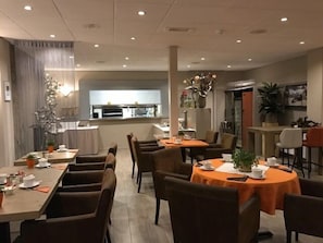 Un espace repas moderne avec plusieurs tables et chaises, une cuisine en arrière-plan et des plantes décoratives.