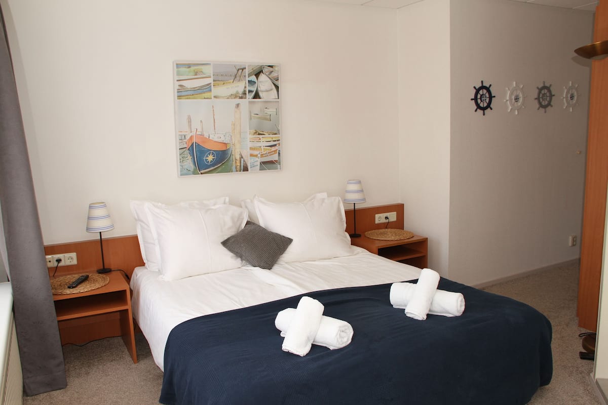 A neatly made bed with white and navy blue bedding, two bedside tables with lamps, a wall-mounted artwork depicting boats, and a set of wall-mounted ship wheels.