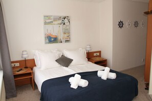 A neatly made bed with white and navy blue bedding, two bedside tables with lamps, a wall-mounted artwork depicting boats, and a set of wall-mounted ship wheels.