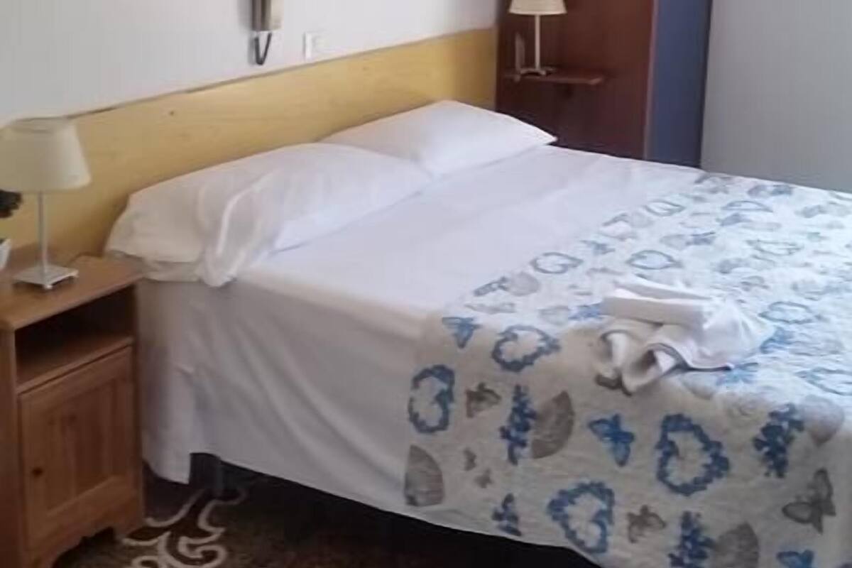 Un dormitorio con cama, una mesita de noche, una lámpara, un armario, un cuadro en la pared y una alfombra estampada.