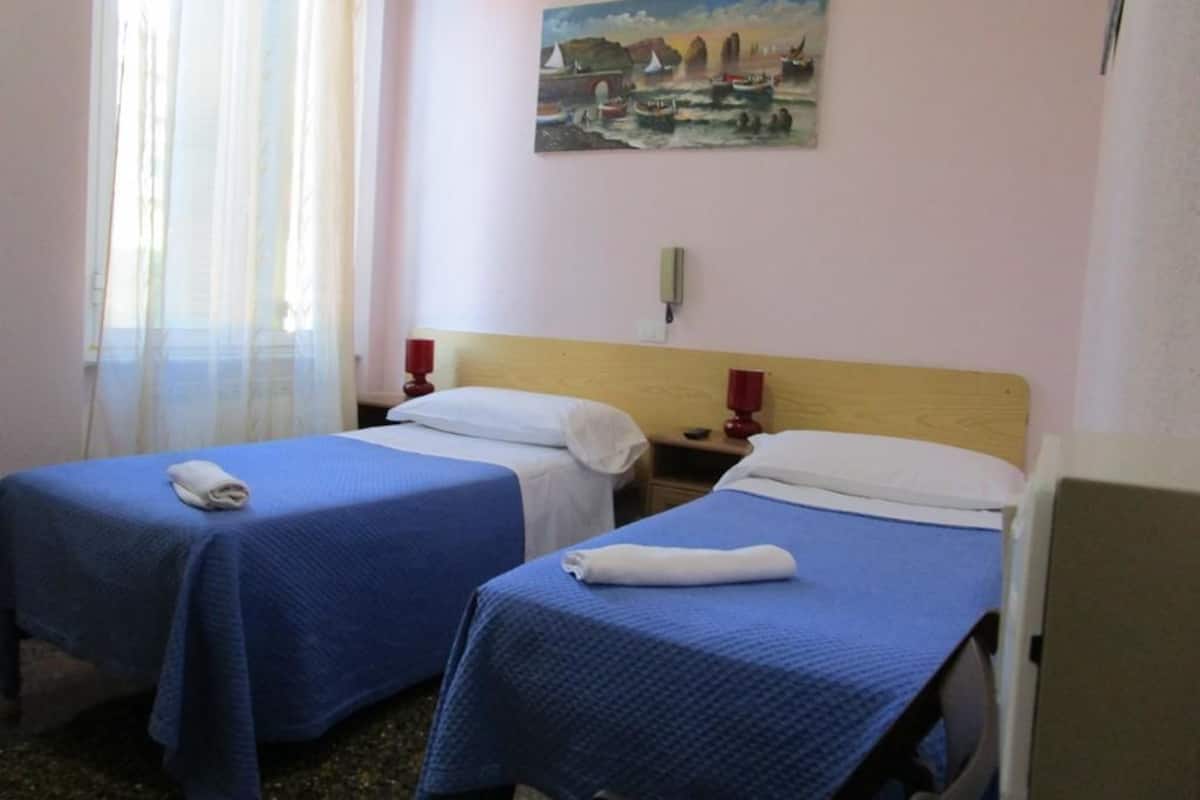 Habitación de hotel con dos camas, cada una con cubrecama azul y almohadas blancas. En la pared hay un cuadro de una escena costera, y una ventana con cortinas traslúcidas que dejan entrar luz natural.