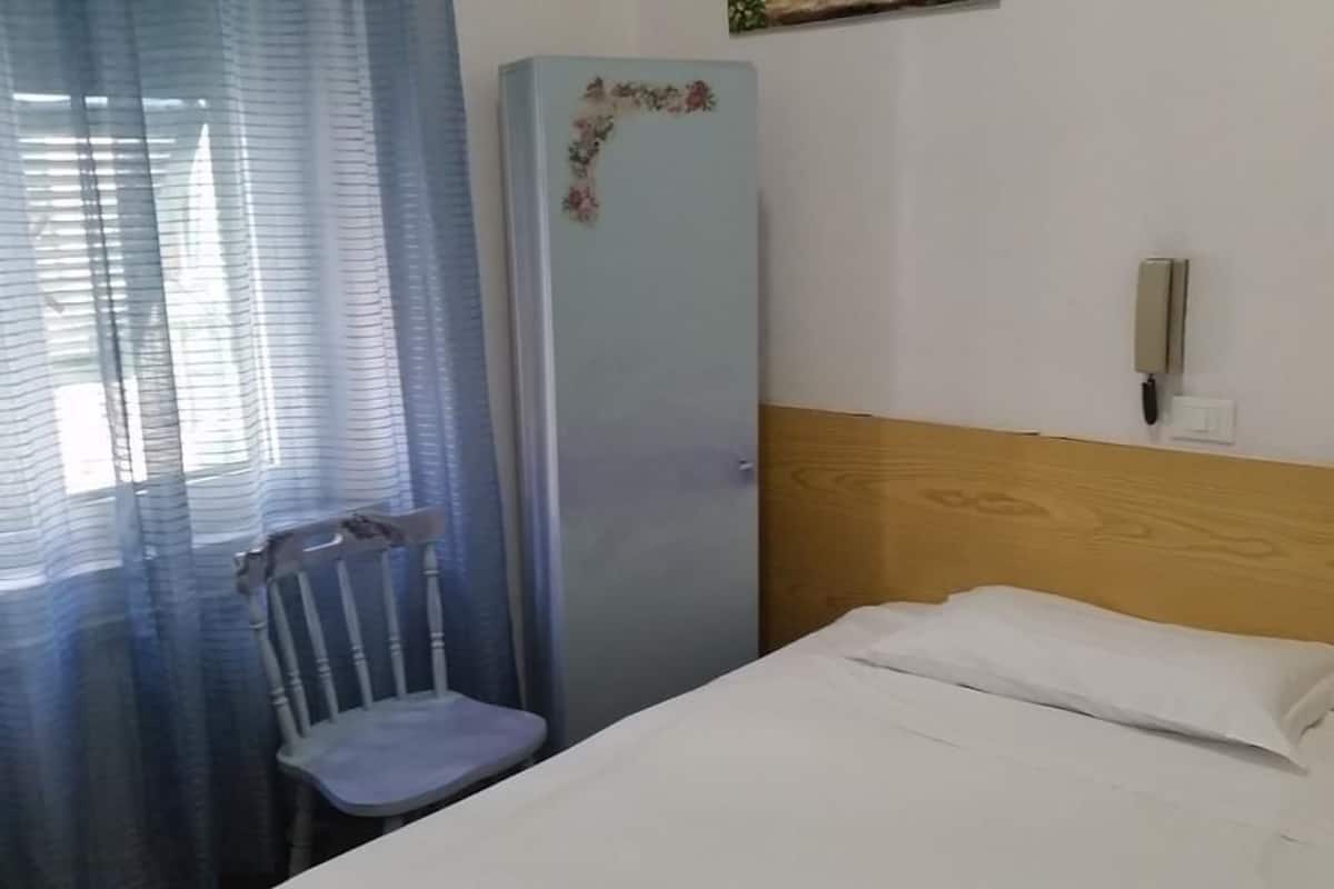 Una habitación pequeña y sencilla con una cama individual, cabecera de madera, una silla y un refrigerador con diseño floral.