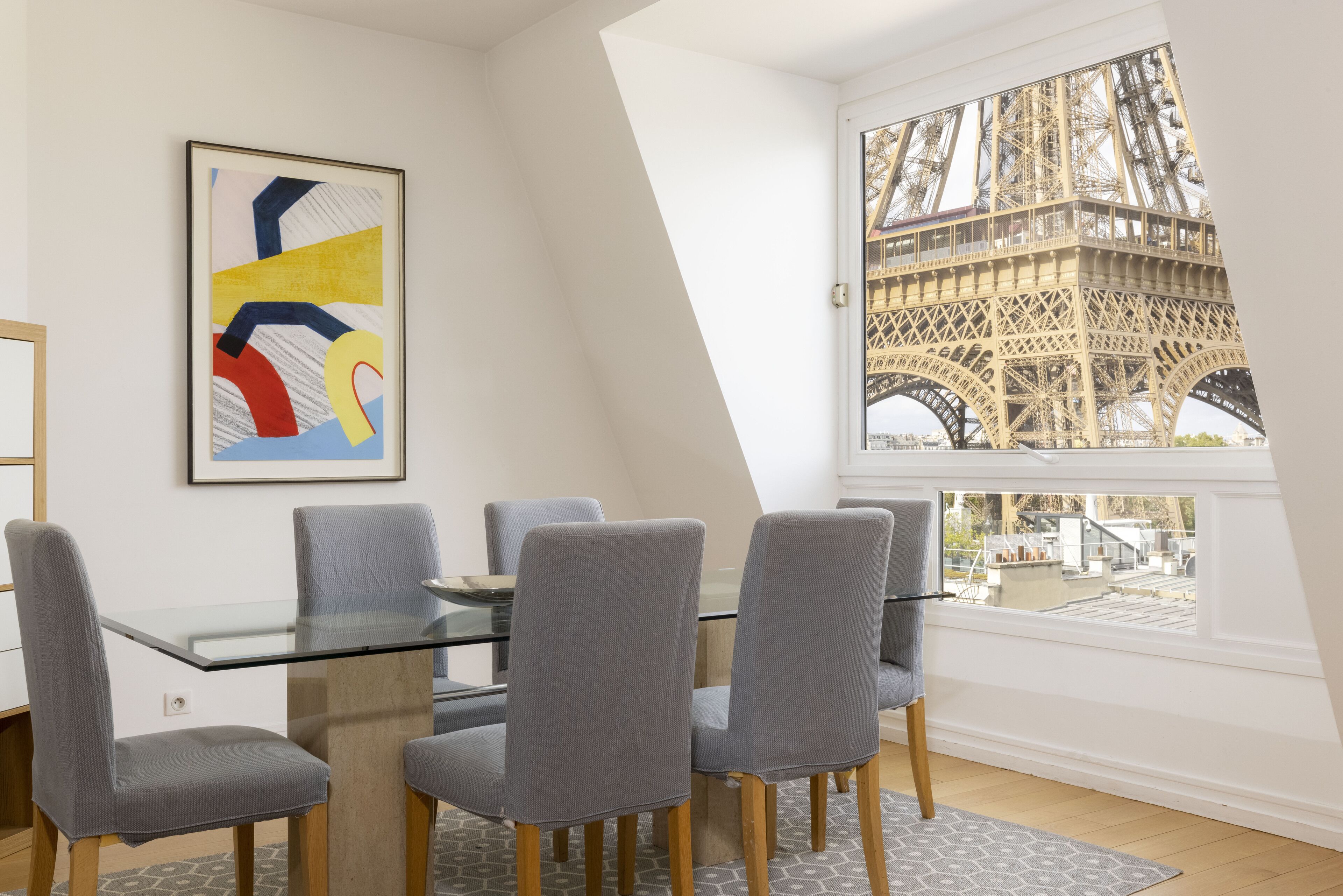 Un coin repas avec une table en verre et des chaises à revêtement gris, une peinture abstraite aux couleurs vives, et une vue sur la Tour Eiffel par une grande fenêtre.