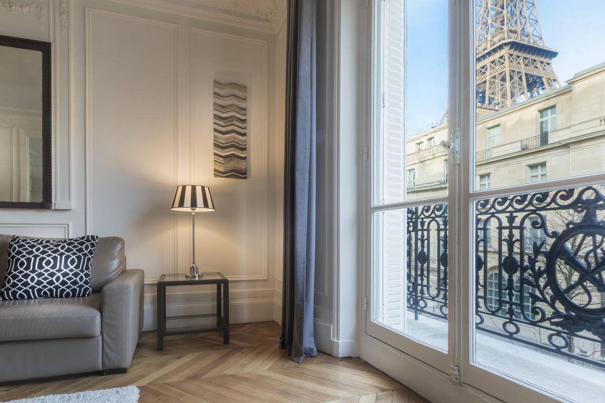 A room with a view of the Eiffel Tower, a sofa with patterned cushions, a floor lamp, and a small table.