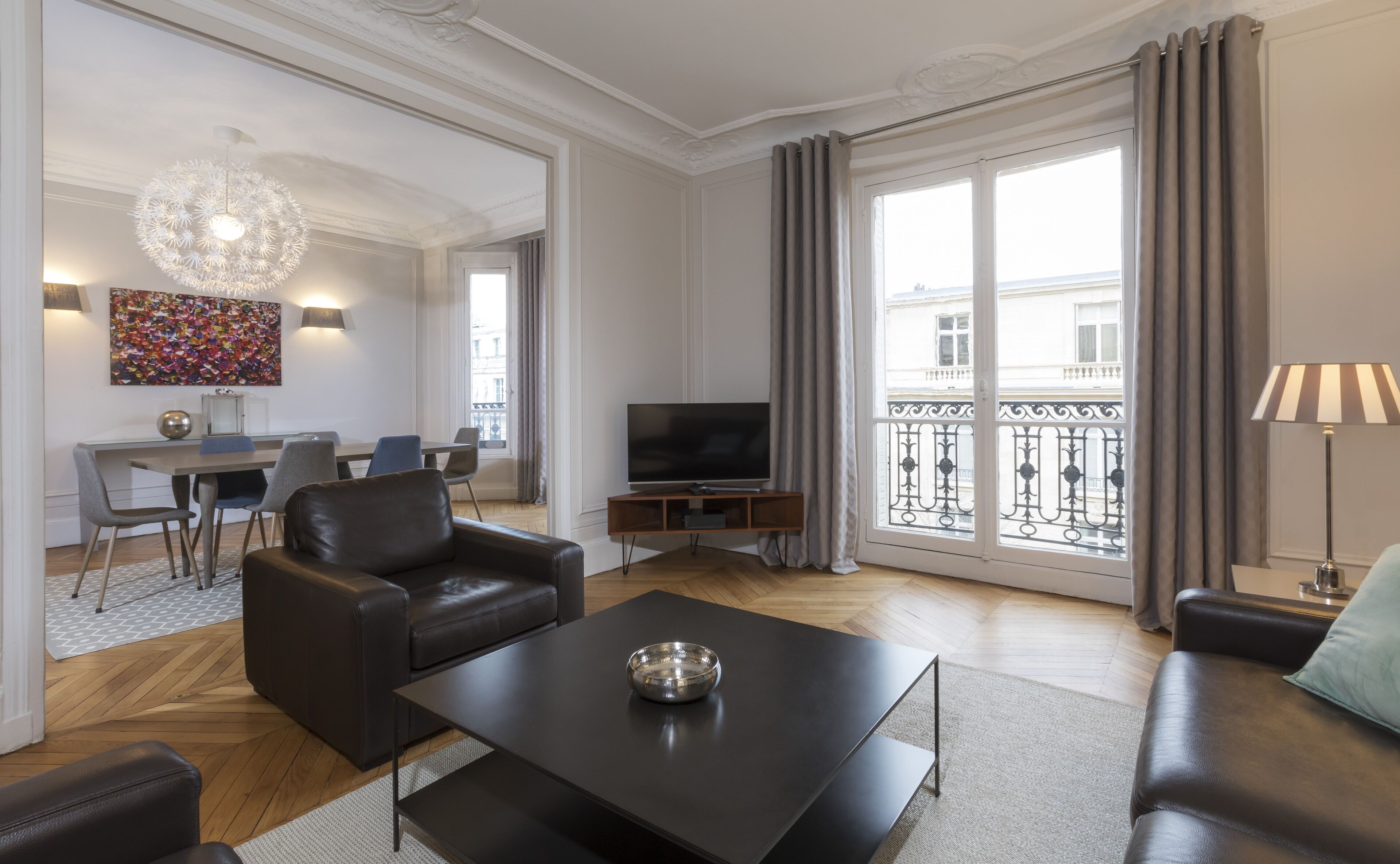 Ein modernes Wohnzimmer mit Flachbildfernseher, Ledersofas, einem Essbereich mit Tisch und Stühlen sowie einem Balkon mit schmiedeeisernem Geländer.