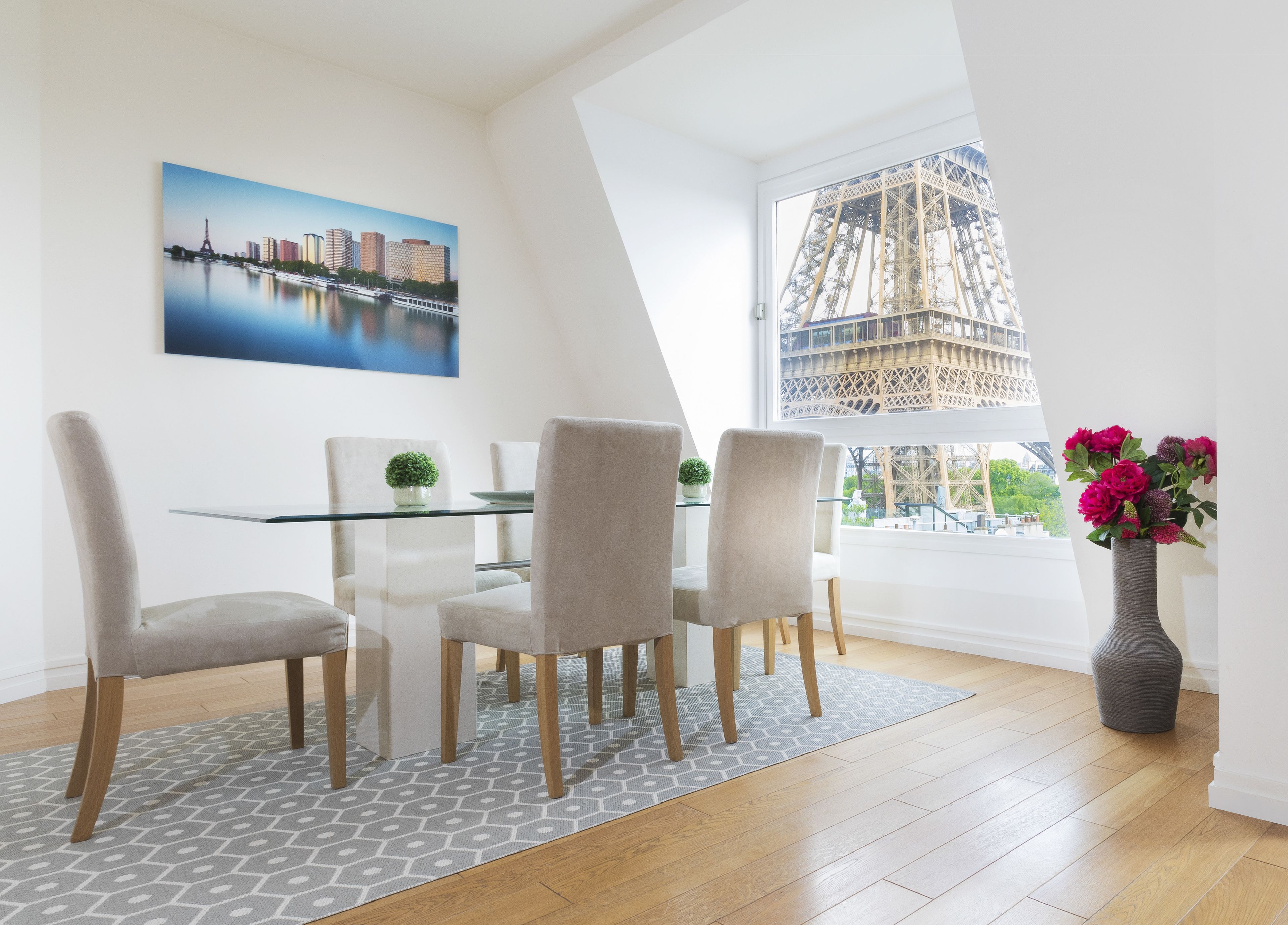 Un espace repas moderne avec une table en verre, quatre chaises, un vase de fleurs et une vue sur la Tour Eiffel par la fenêtre.