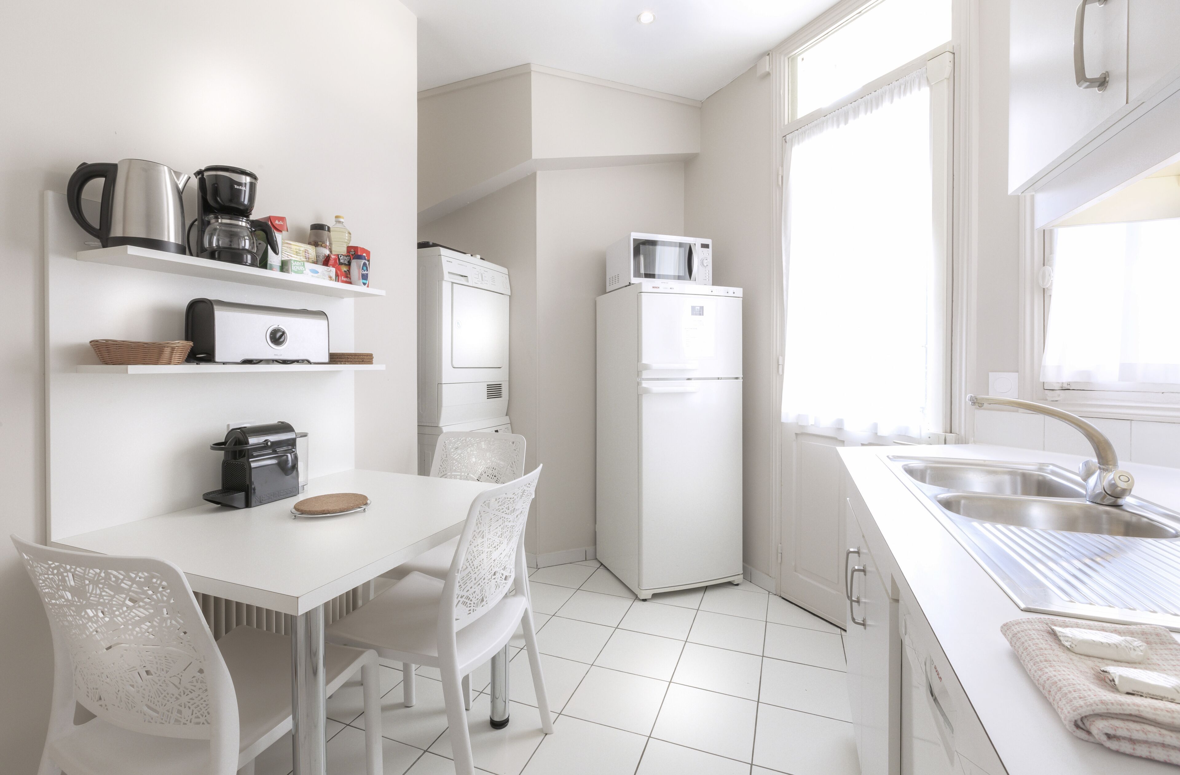 A modern kitchen with white appliances, a dining area with white chairs, and a large window providing natural light.