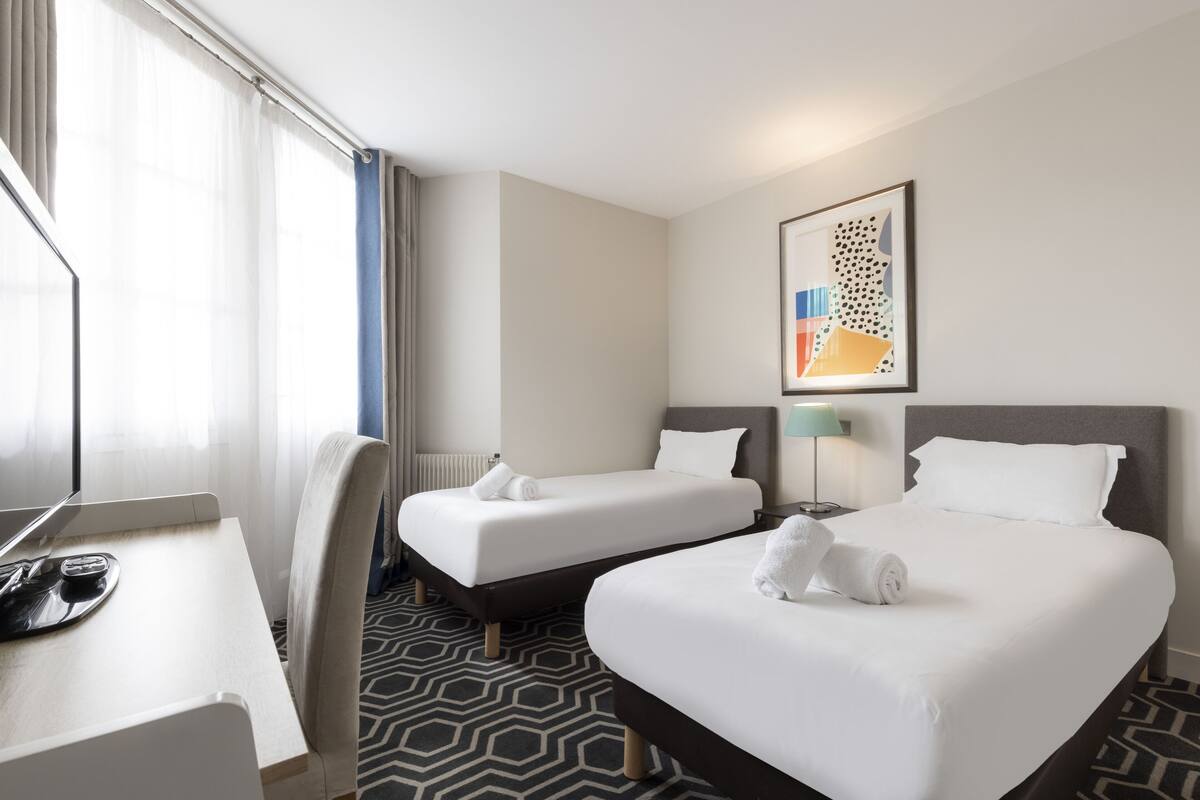 A hotel room with two beds, a desk, a chair, and a lamp.