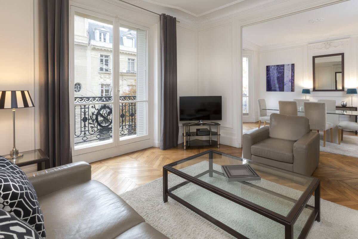 A modern living room with a glass coffee table, a sofa, a TV on a stand, and a dining area with chairs and a table.
