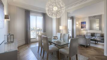 A modern hotel room with a glass dining table, comfortable seating, a chandelier, and a city view.