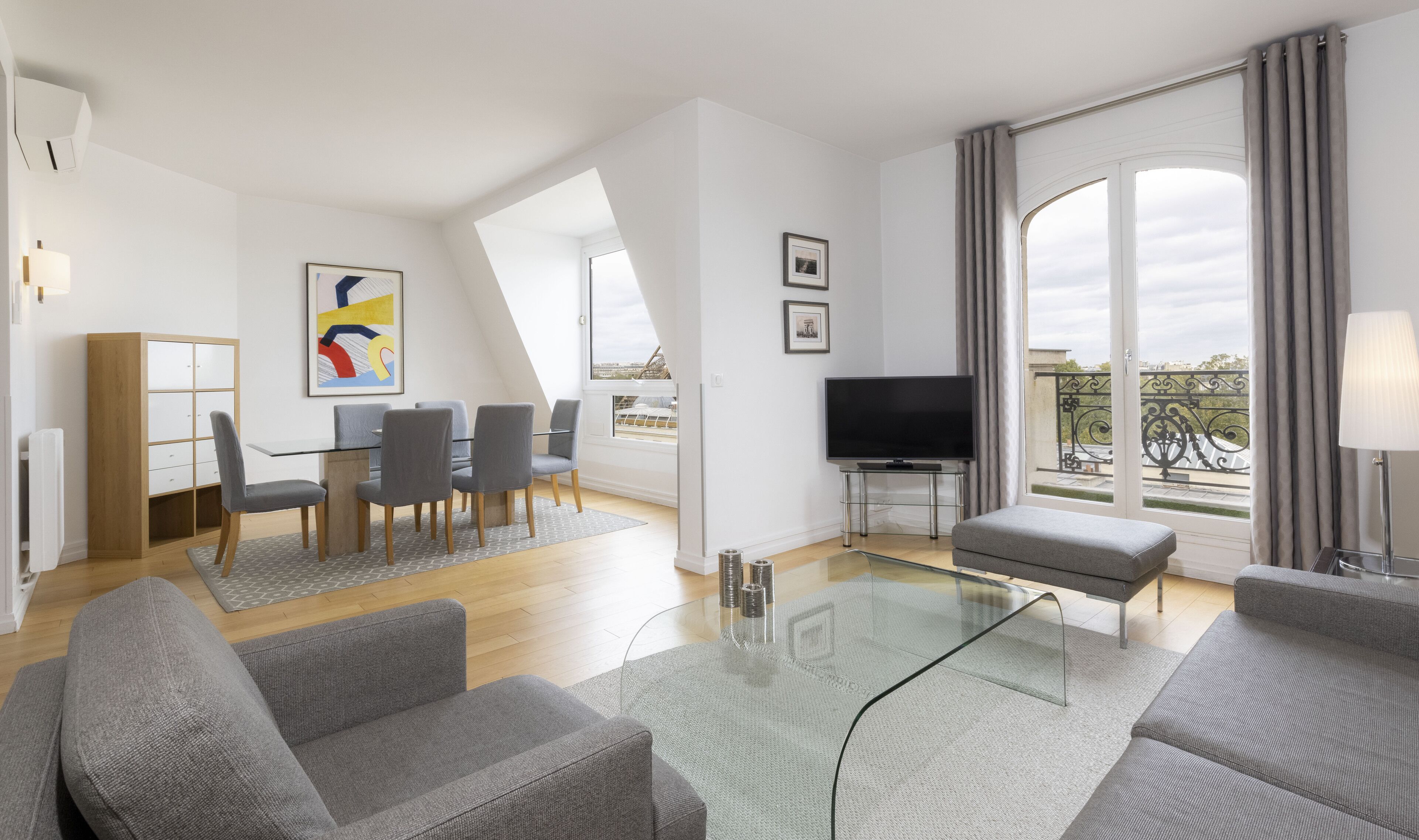 A modern living room with a glass coffee table, grey sofas, a TV, and a dining area with a wooden cabinet and grey chairs.