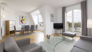 A modern living room with a glass coffee table, grey sofas, a TV, and a dining area with a wooden cabinet and grey chairs.