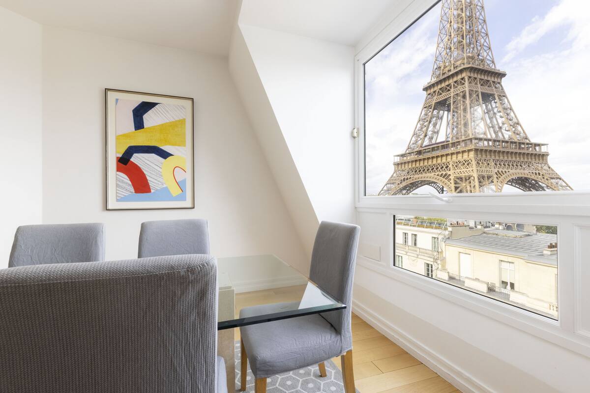 A room with a glass table, two grey chairs, a framed abstract painting, and a view of the Eiffel Tower through a large window.