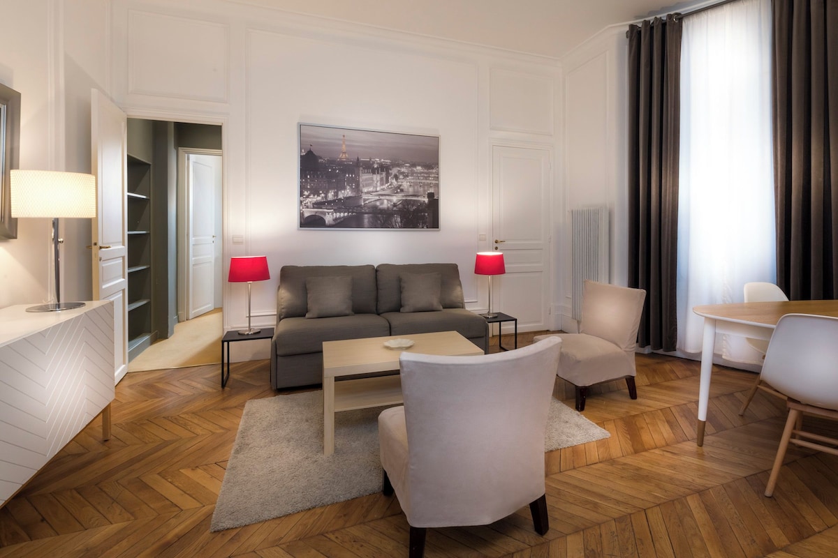 Un salon moderne avec un canapé, deux fauteuils, une table basse et une table à manger avec des chaises. Un tableau est accroché au mur, une lampe se trouve de chaque côté du canapé, et la fenêtre offre une vue sur la ville.