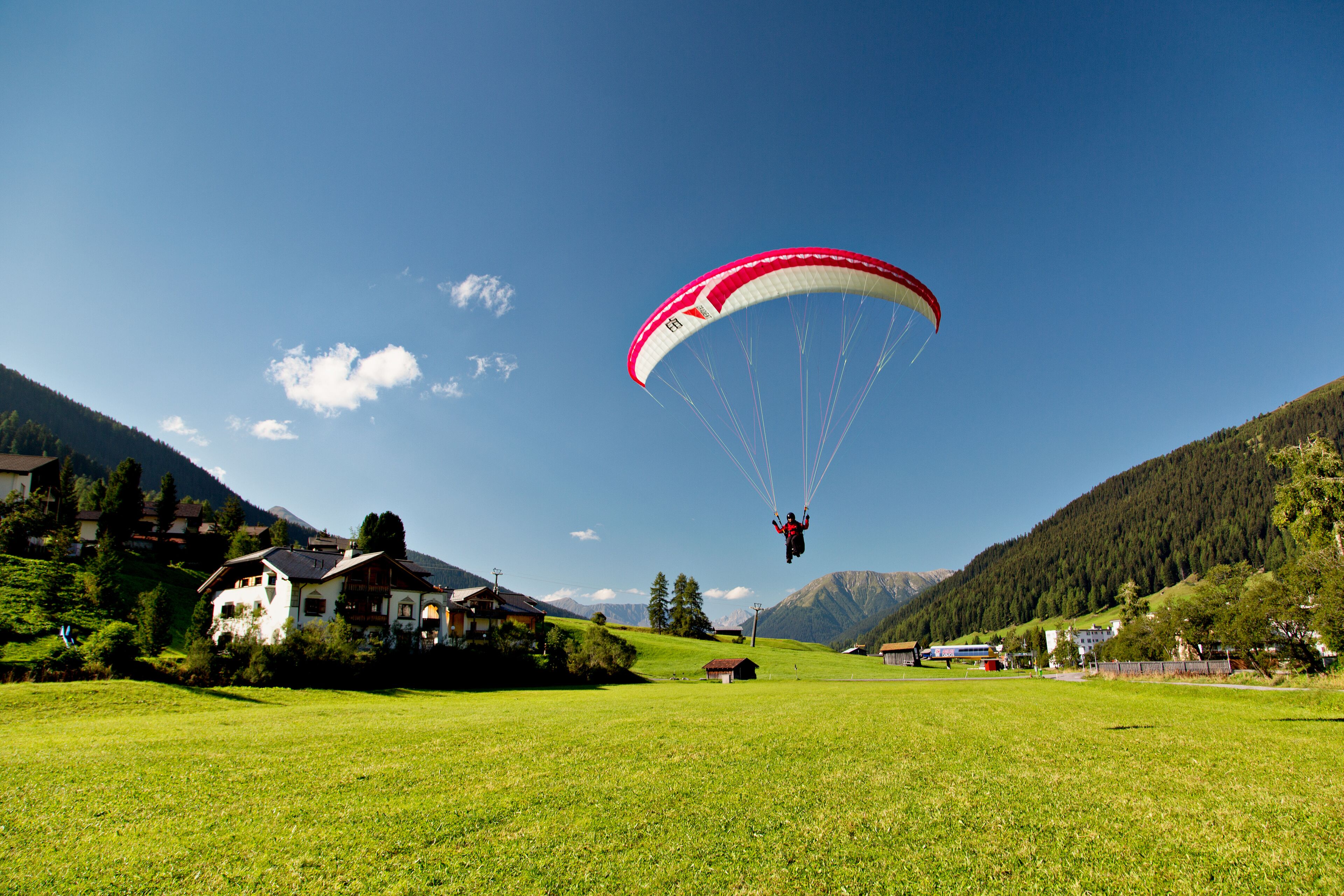 A person paragliding over a green field with houses and mountains in the background.