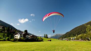 A person paragliding over a green field with houses and mountains in the background.
