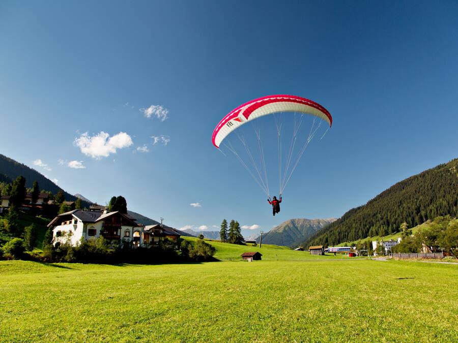 A person paragliding over a green field with houses and mountains in the background.