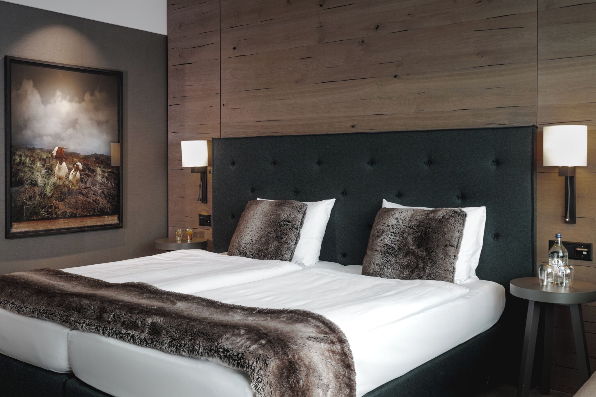 Habitación de hotel con una cama grande, cabecera, dos lámparas de noche, mesitas de noche, un cuadro enmarcado en la pared y una manta de piel sobre la cama.